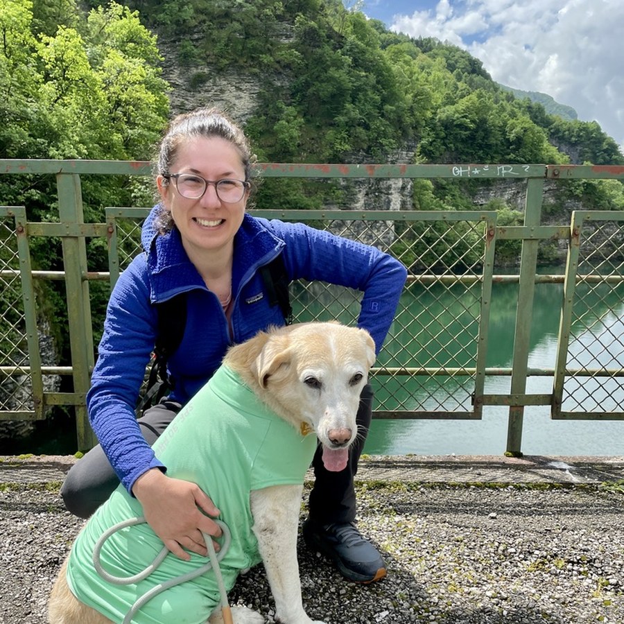 Lavinia crouched beside Sara, a yellow Labrador in a green coat, on a path beside a lake
